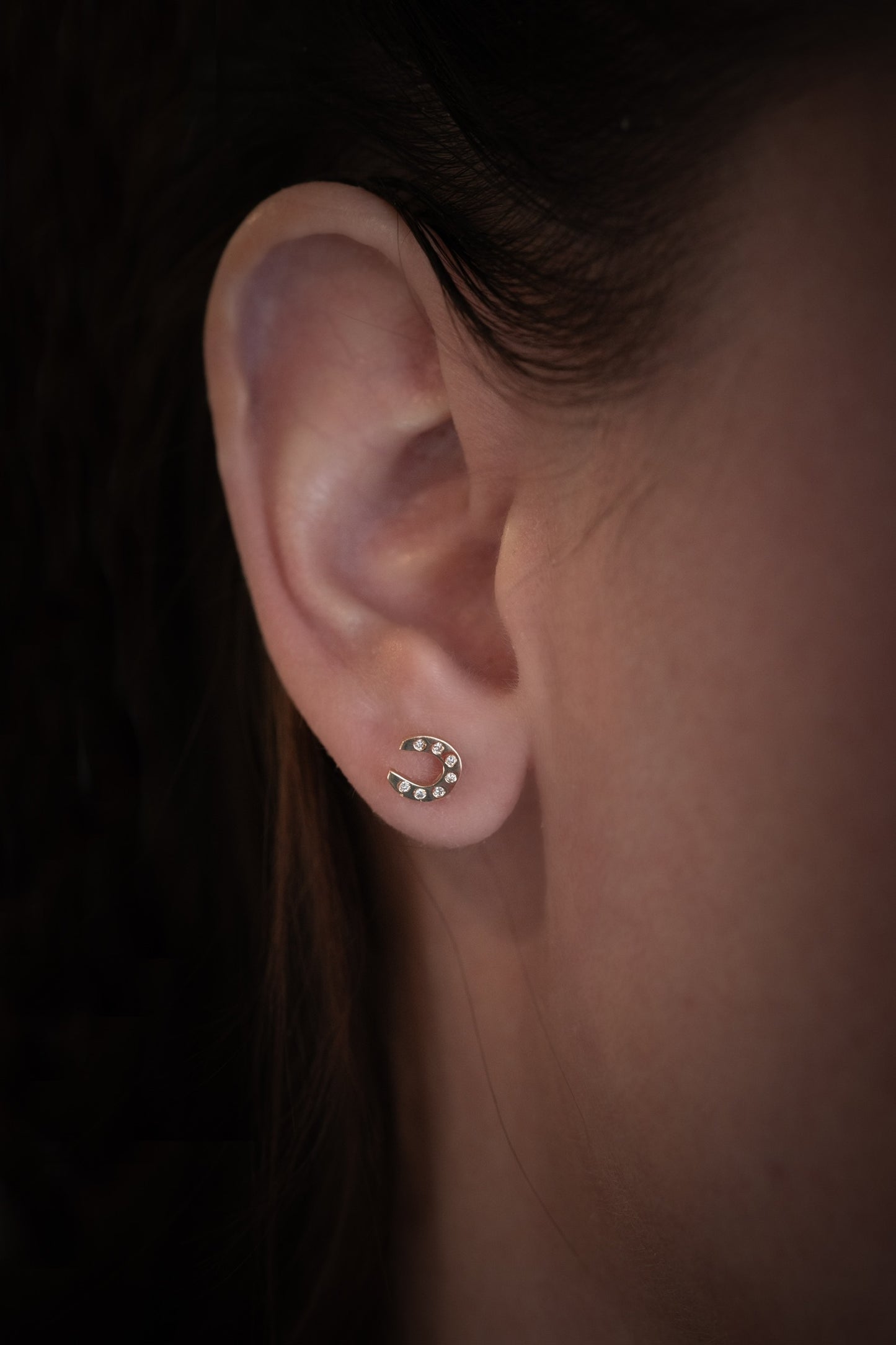 Close-up of an ear wearing solid gold horseshoe lab diamond stud earring with a dark background